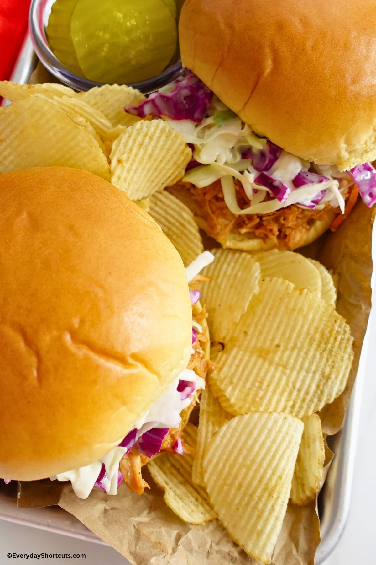 two barbeque pulled chicken sandwiches on a tray with chips and pickles