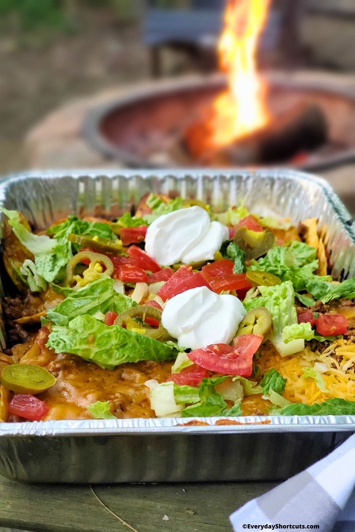 nachos with toppings in a foil pan next to a campfire