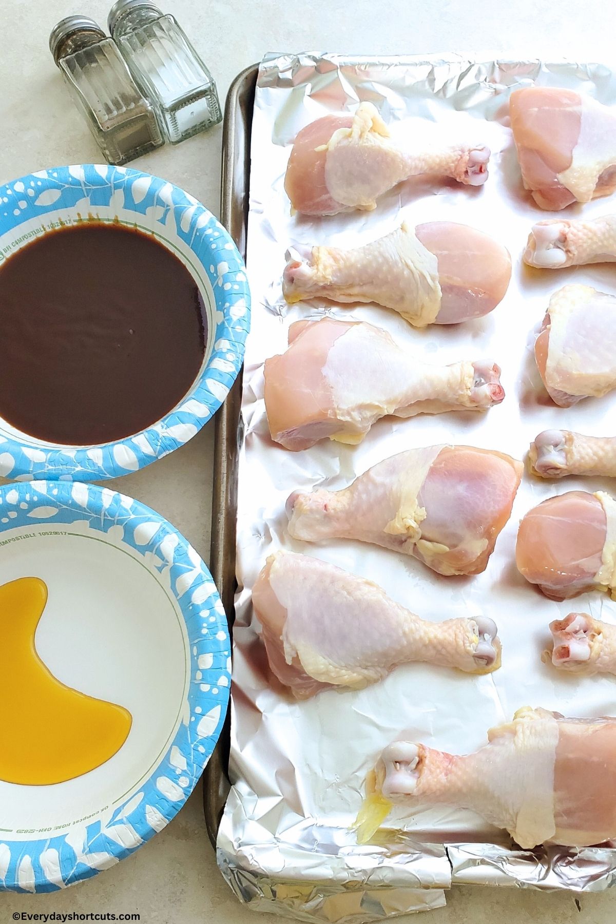 raw chicken on a baking sheet with foil salt and pepper shakers bbq sauce in a bowl and maple syrup in a bowl