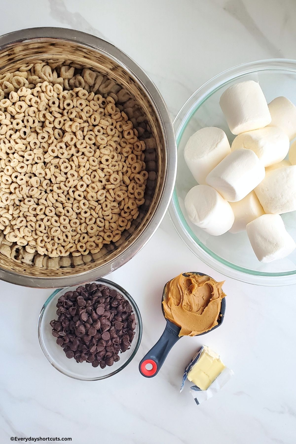 Cheerios marshmallows and chocolate chips in separate bowls peanut butter in a measuring cup and butter on a white board