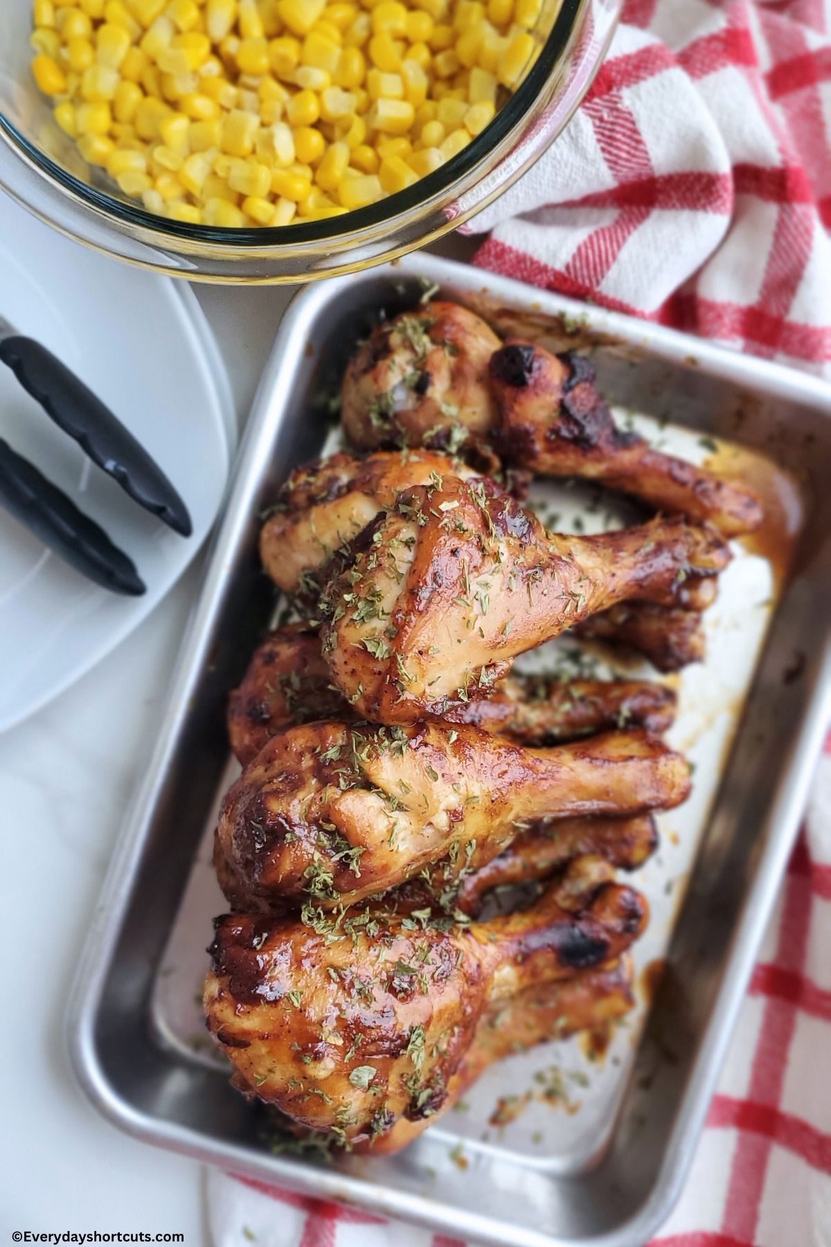 barbeque chicken drumsticks on a baking sheet with corn in a bowl next to white plates