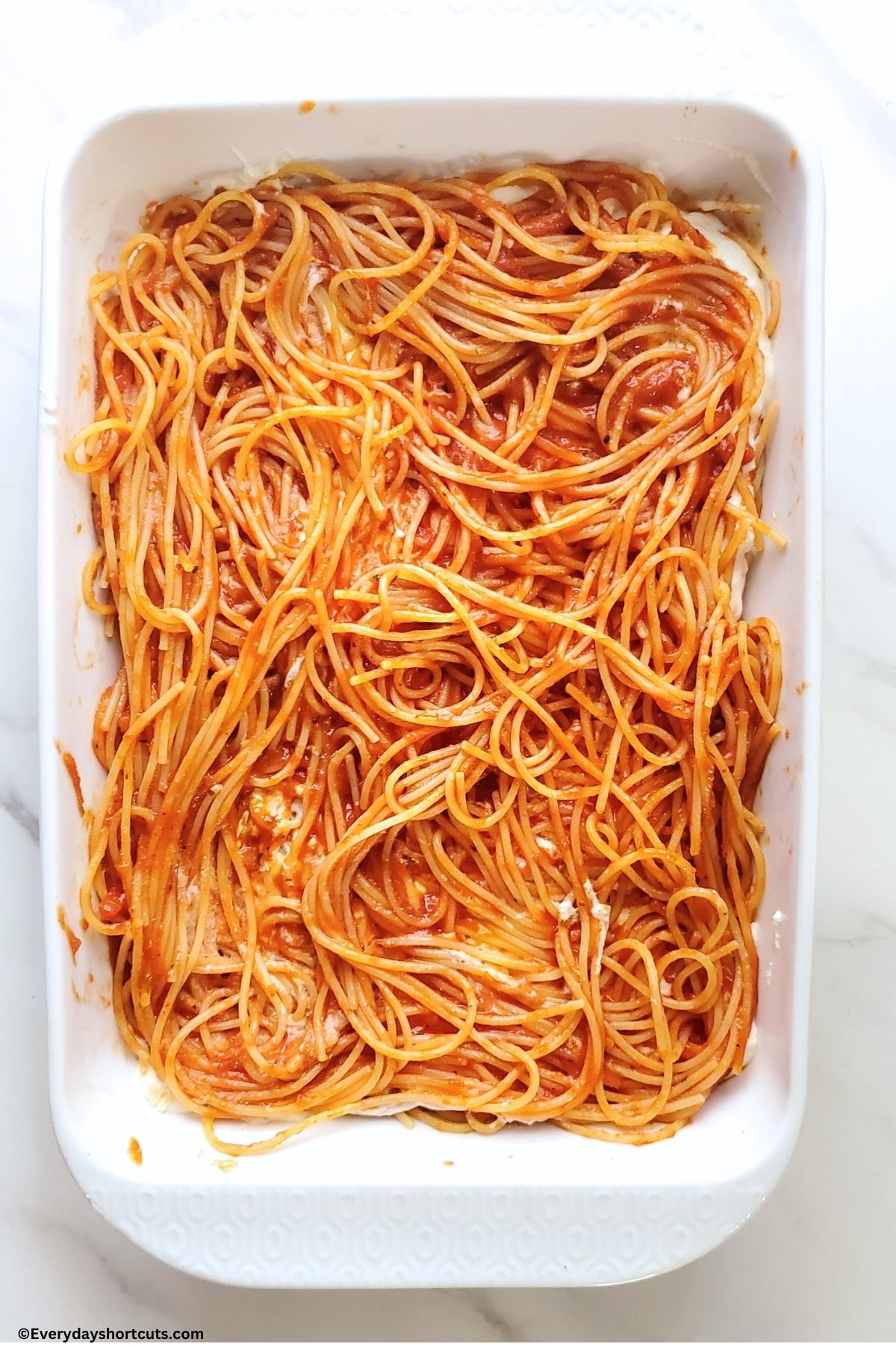 cooked spaghetti noodles with marinara sauce on top of creamy cheese mixture in white baking dish