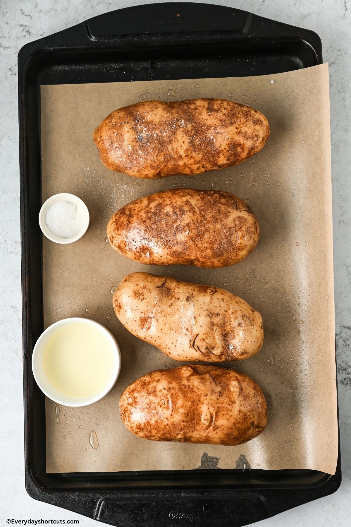 four russet potatoes on a lined baking sheet next a bowl of salt and oil