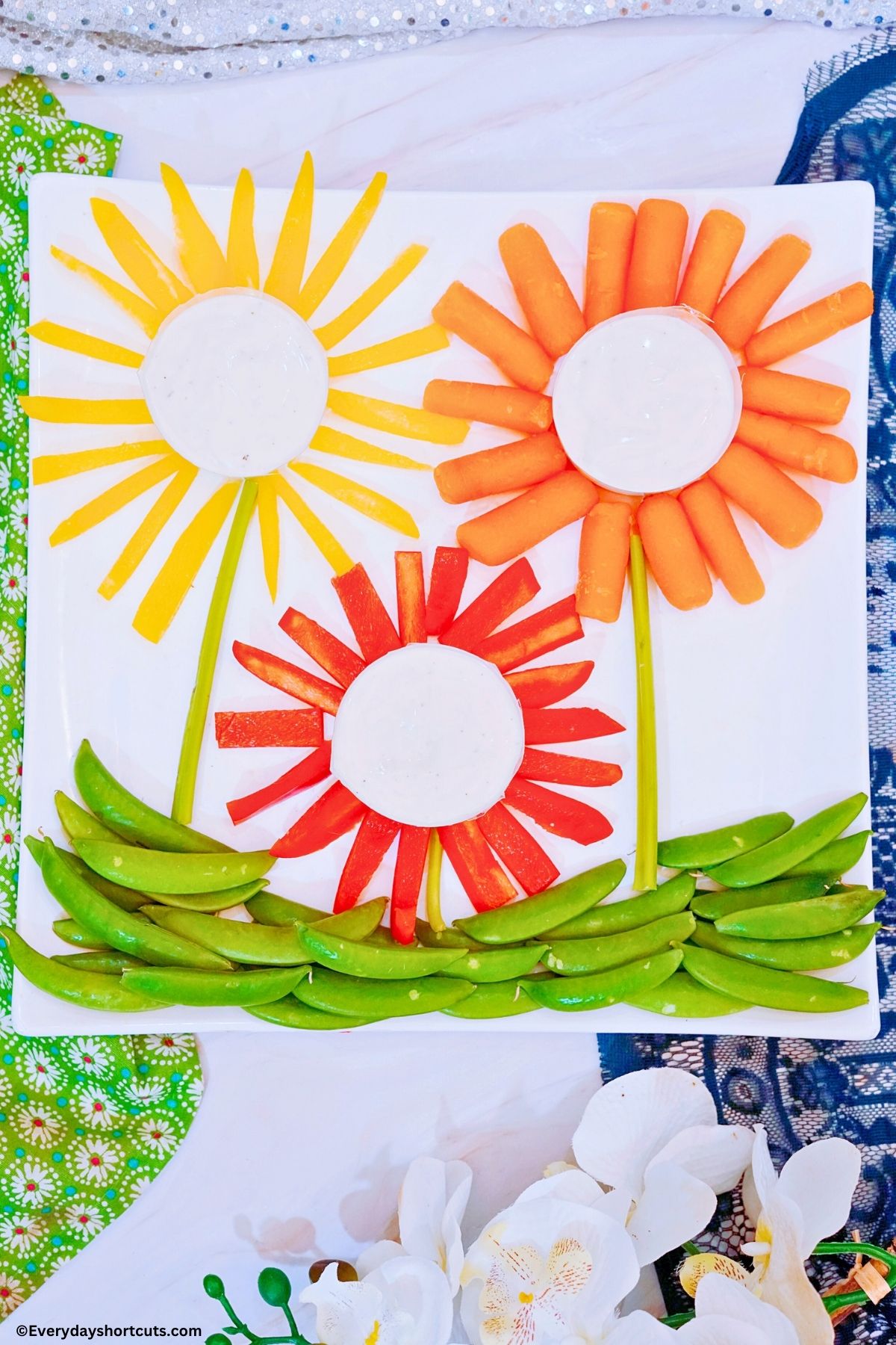 carrots red and yellow bell peppers in the shape of flowers with bowls of ranch dressing in the middle of each one celery sticks for stems and snap peas for grass on a platter