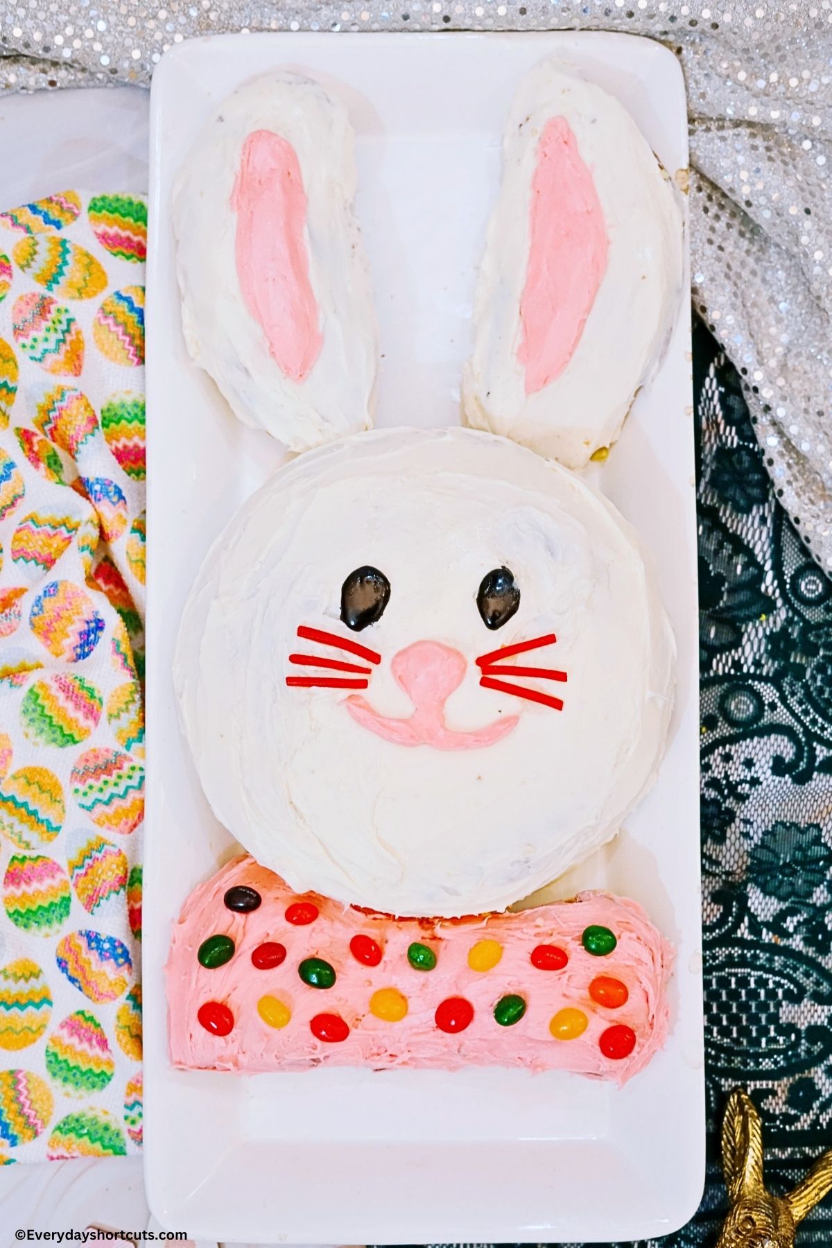 Bunny Shaped Cake on a white platter frosted in white and pink icing and decorated with jelly beans for the bow tie, black icing, Twizzlers, and pink frosting for the bunny face