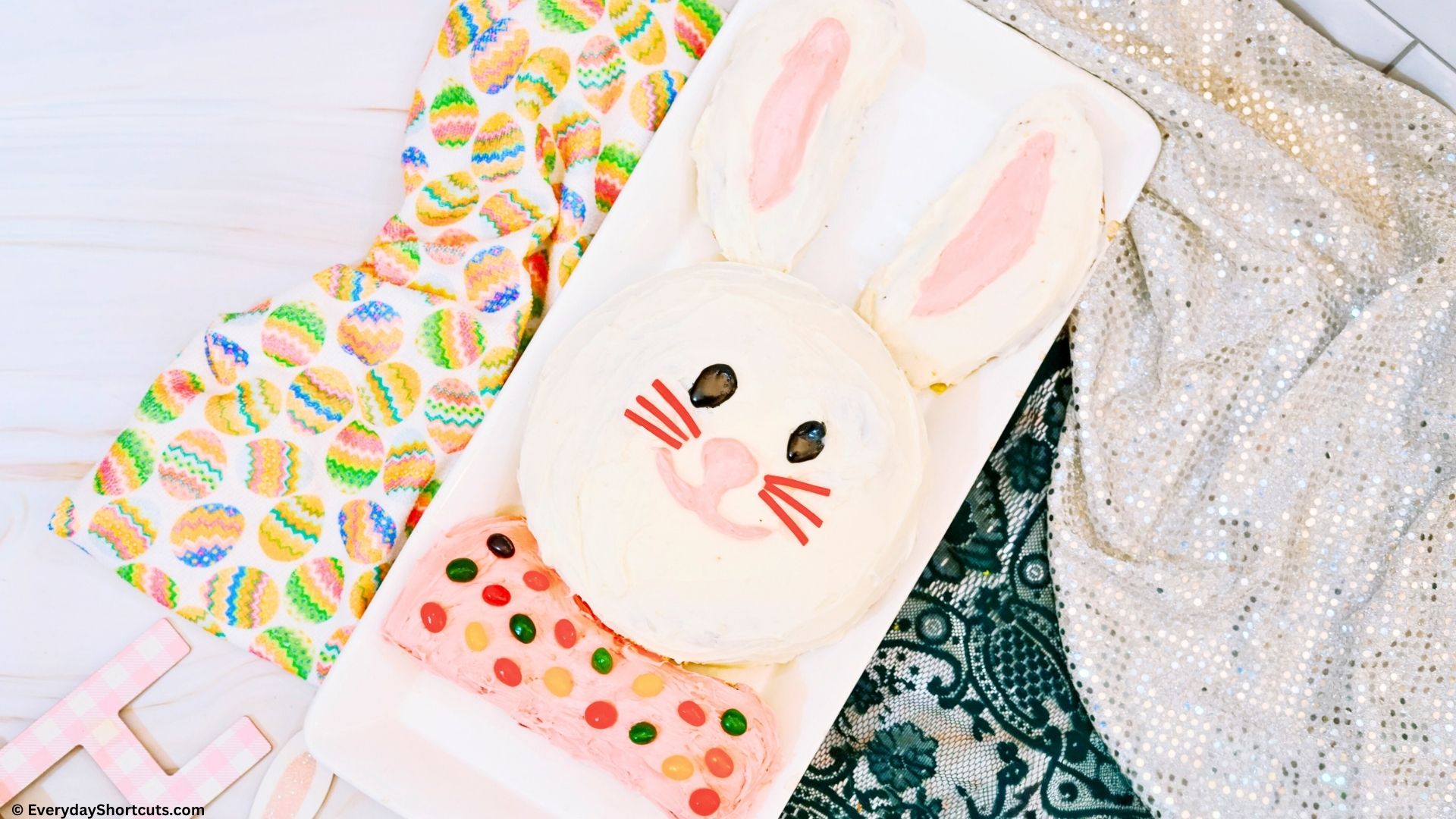 Easter Bunny Cake on a white platter frosted with white and pink icing and decorated with black icing pink frosting twizzlers to make the bunny face and jelly beans for the bow tie