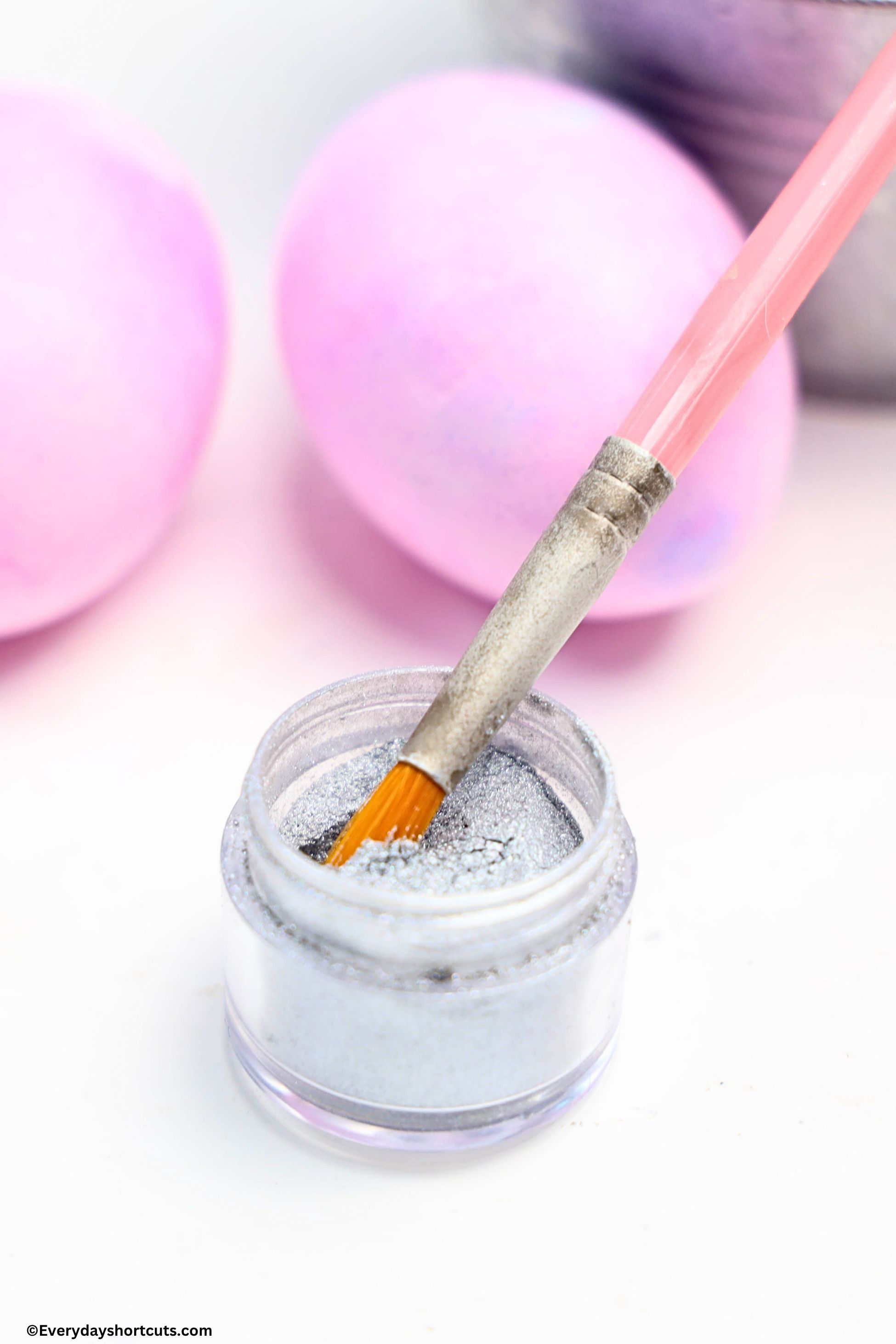 edible silver glitter dust with a paint brush in it next to two pink colored eggs