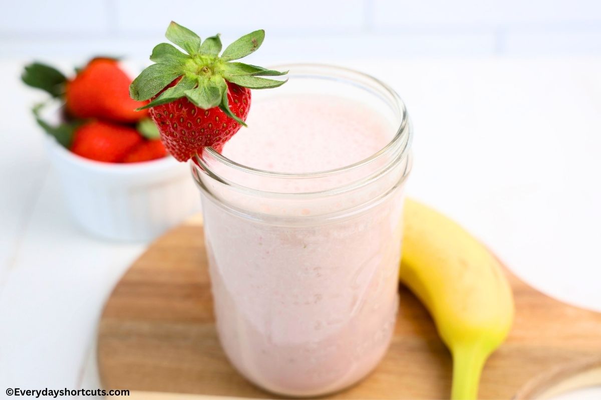 strawberry banana smoothie in a glass with a strawberry garnish on a wooden board next to a banana and a bowl of strawberries