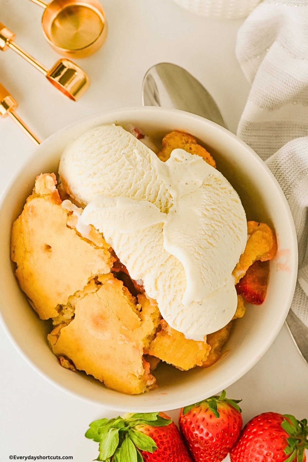 bowl of strawberry cobbler with vanilla ice cream on top next to strawberries and a spoon
