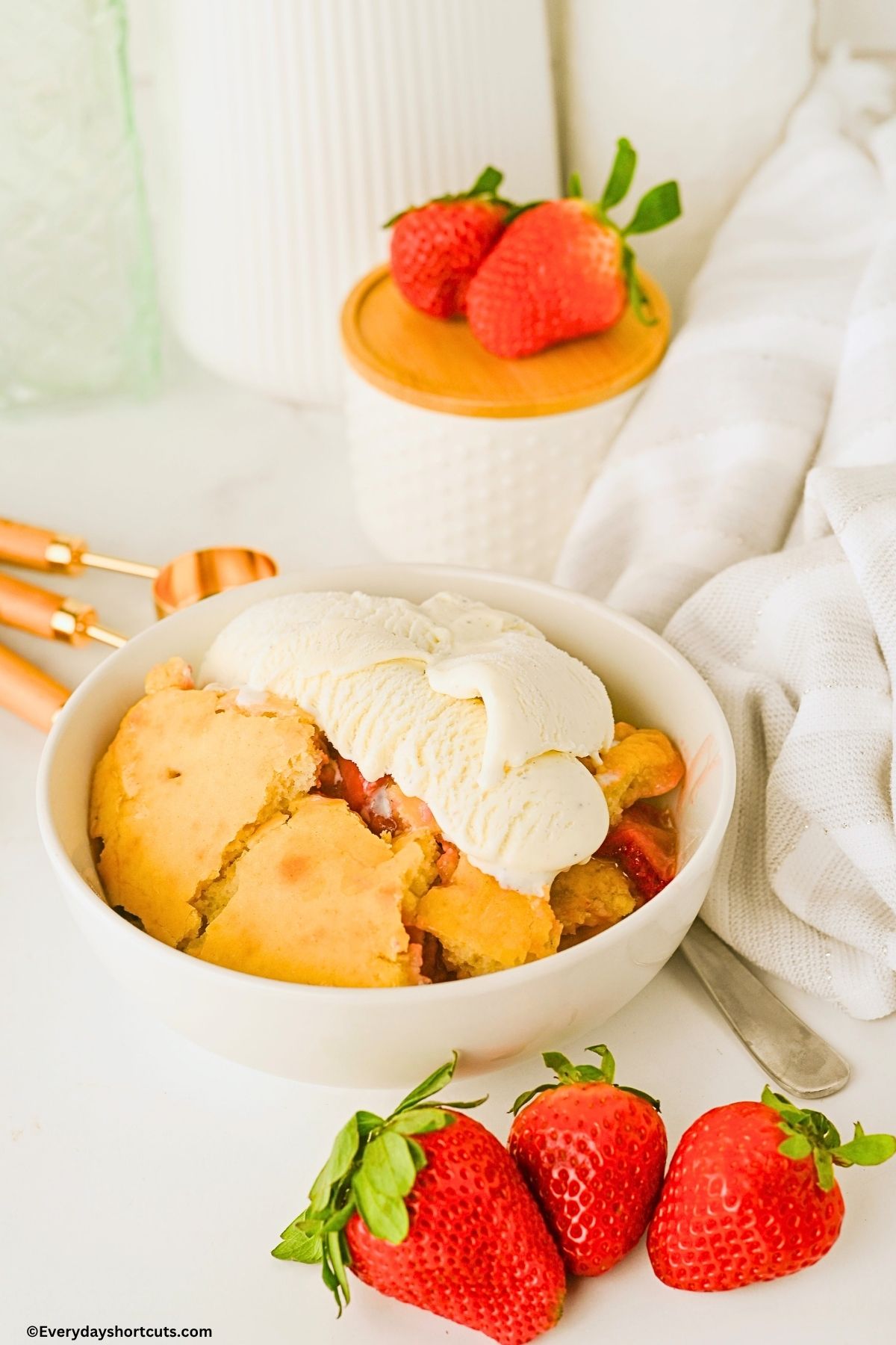 strawberry cobbler with ice cream in a bowl next to measuring spoons, strawberries and a spoon