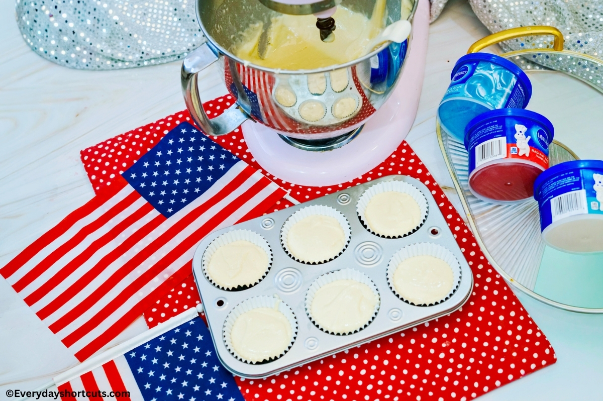 cake mix batter in cupcake liners in a muffin tin next to red white and blue frosting containers