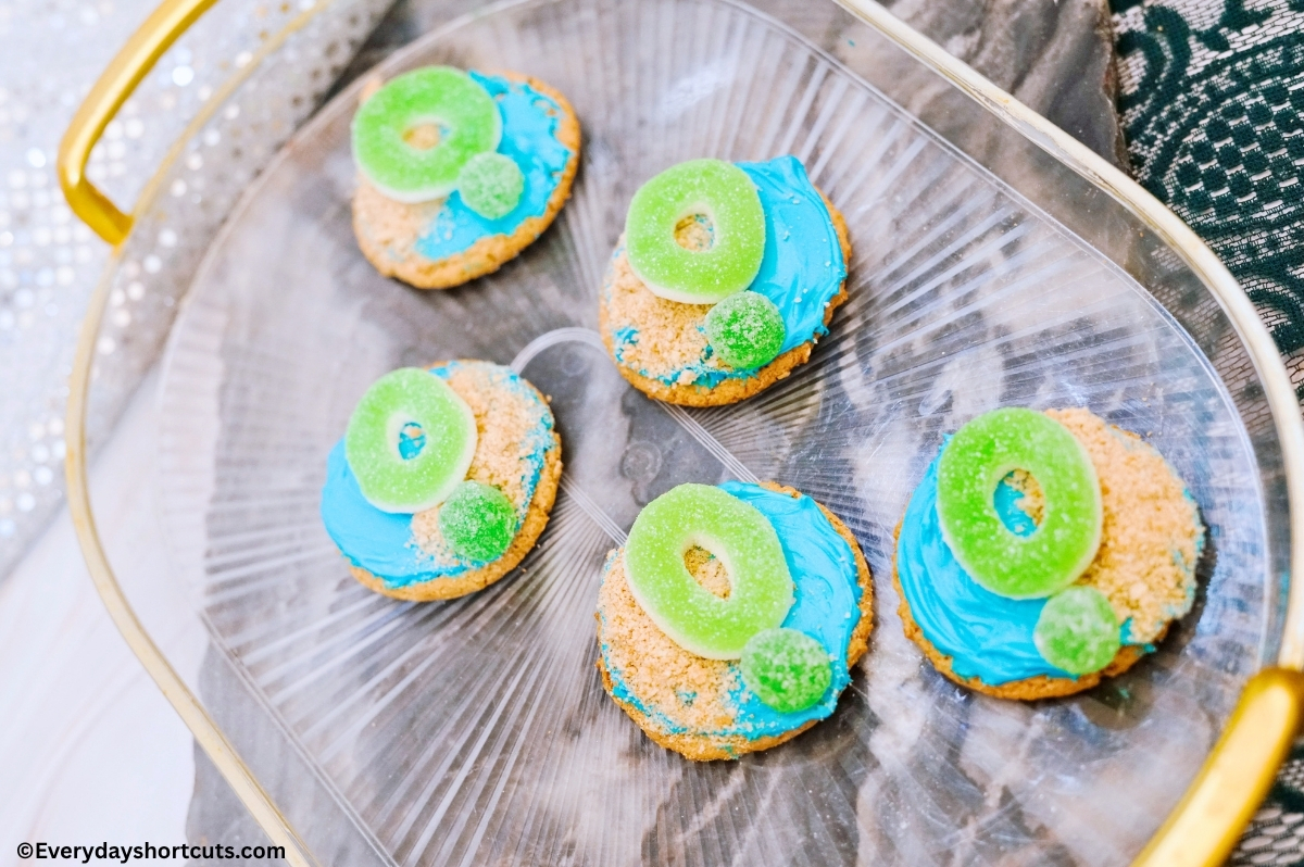 sugar cookies decorated with blue frosting, crushed graham crackers to look like sand, one green gummy ring and one green gumdrop on each cookie