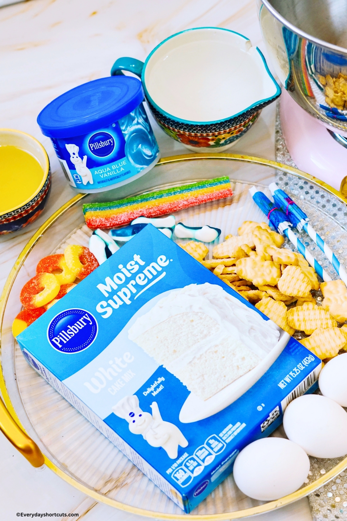 ingredients shown: white cake mix, teddy grahams, rainbow candy strips, gummy sharks, gummy peach rings, paper umbrellas, blue frosting, eggs, water and oil