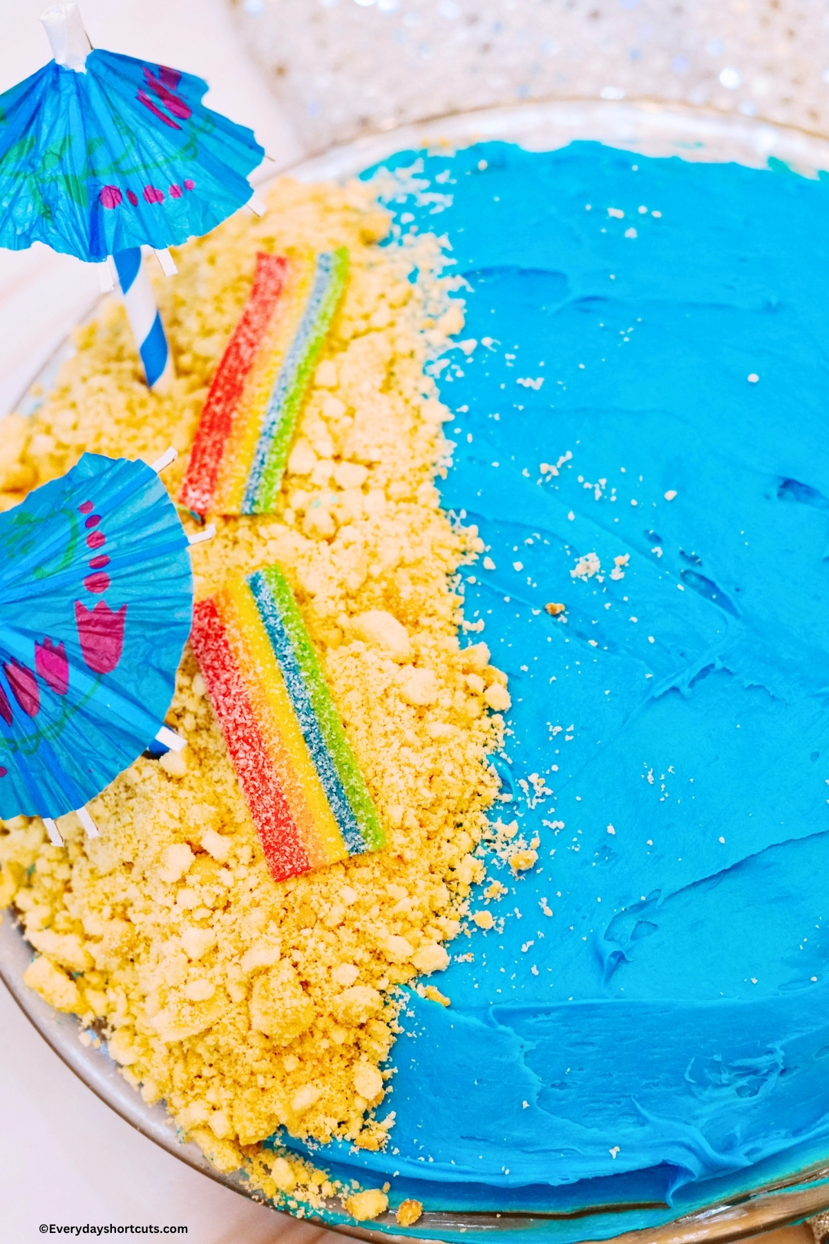 blue frosted cake with crushed teddy grahams on one half with rainbow candy strips as beach towels and paper umbrellas