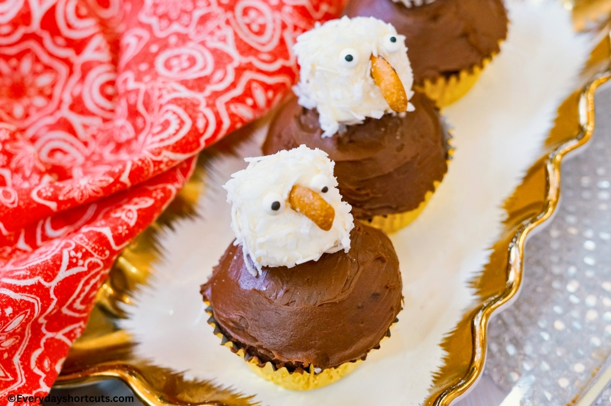 chocolate cupcakes with a coconut coated marshmallow on top, candy eyes and a cashew beak to make it look like a bald eagle on a white and gold tray