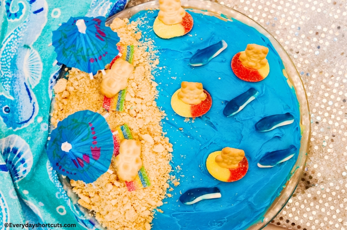 a blue frosted round cake decorated to look like the beach with edible sand, gummy sharks, peach rings for innertubes and teddy grahams for people