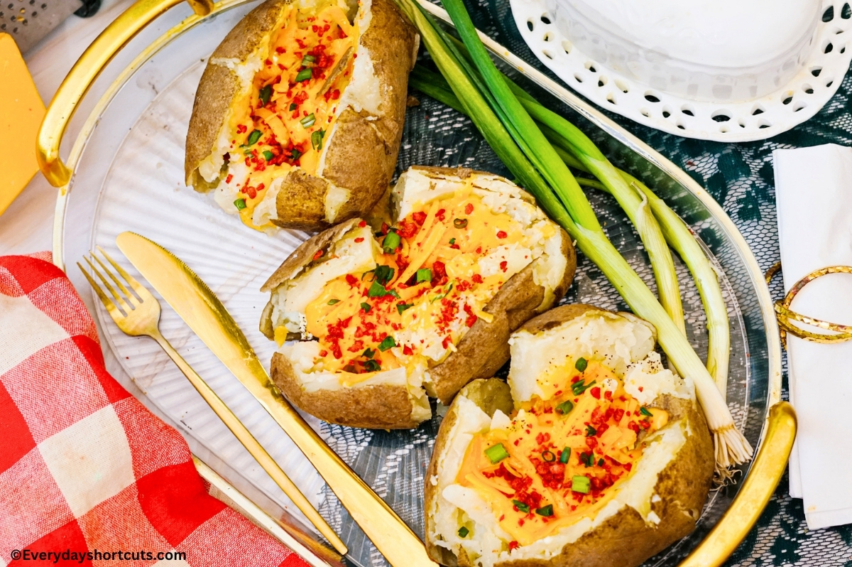3 baked potatoes with cheese, bacon bits and green onions on a clear tray next to a red and white napkin, fork and knife