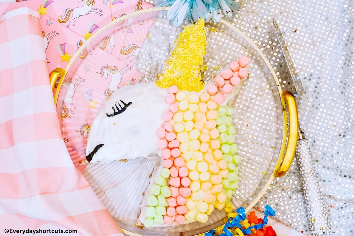 cake shaped in a unicorn and decorated on a clear tray next to a pink and white checkered napkin