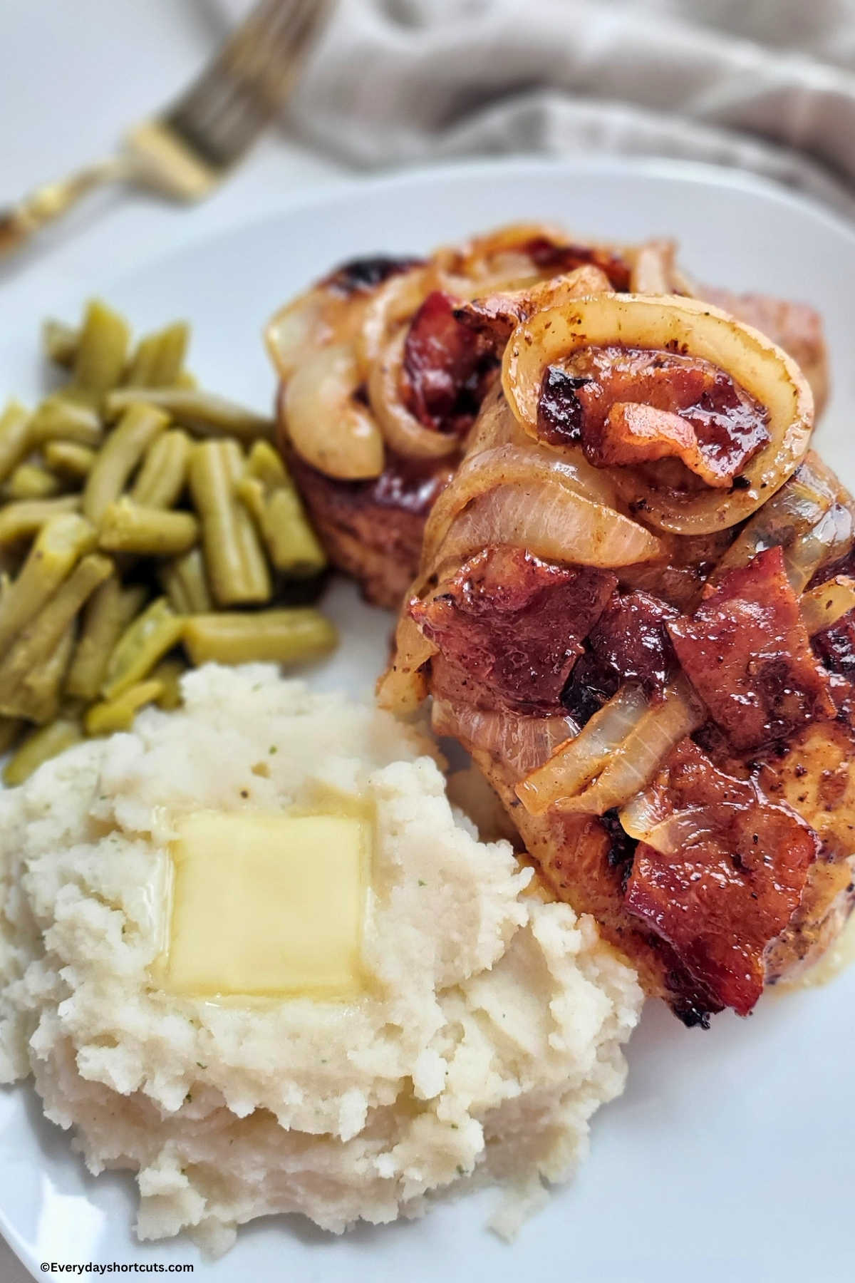 caramelized onions and bacon on top of pork chops next to mashed potatoes and green beans