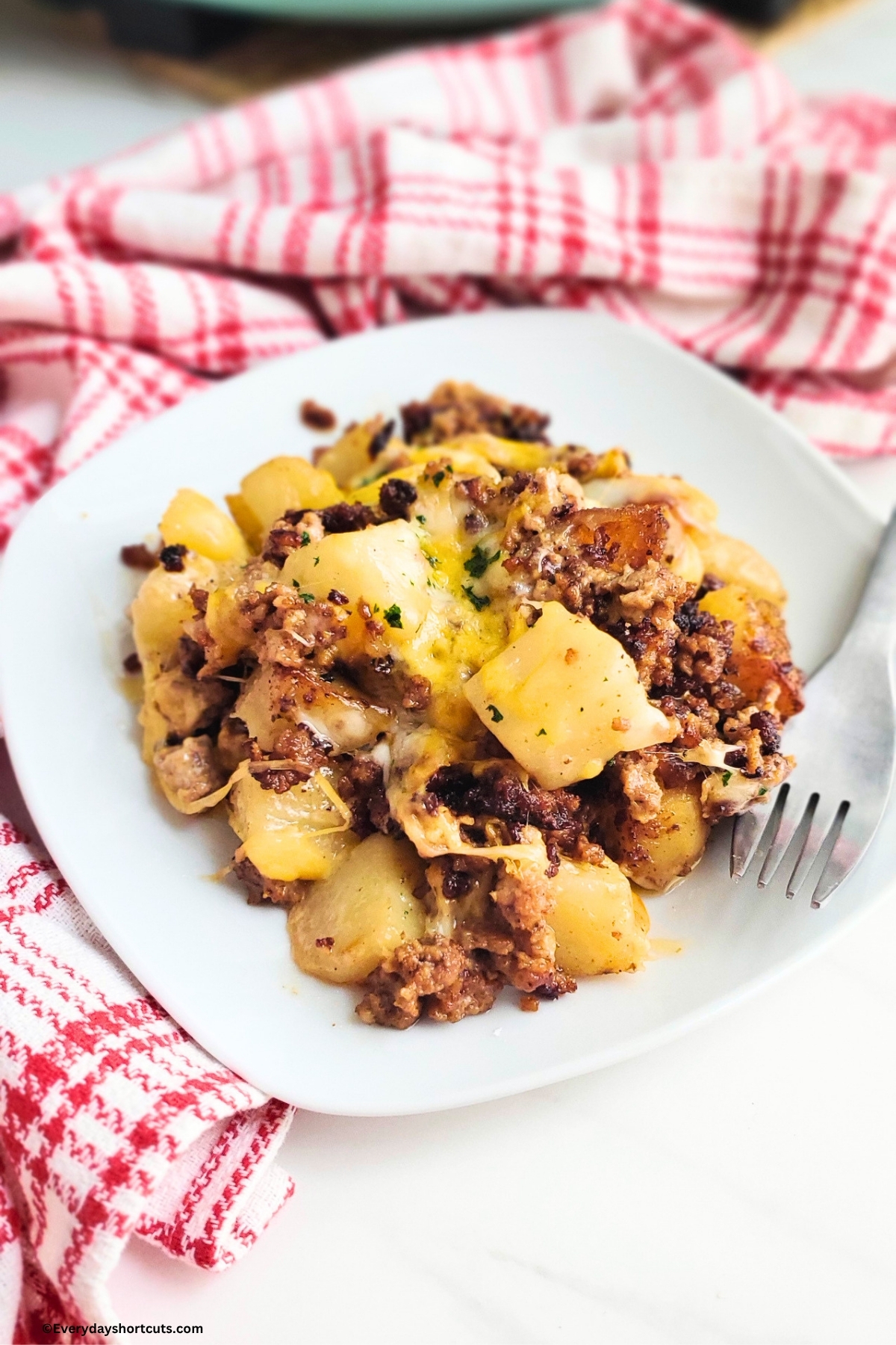 cheesy ground beef and potatoes on a white plate next to a fork and red and white napkin