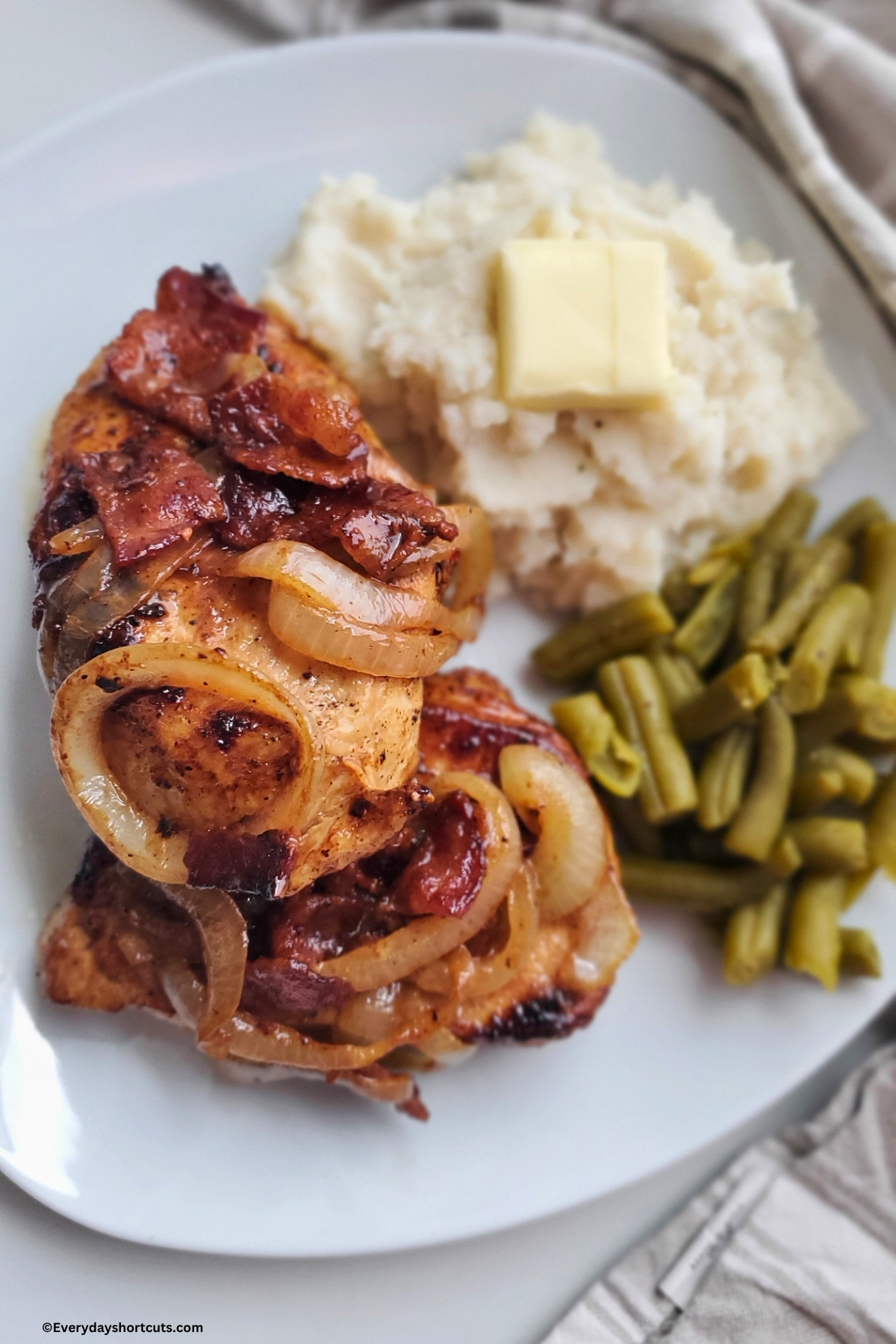 bacon and caramelized onions on top of pork chops on a plate with green beans and mashed potatoes