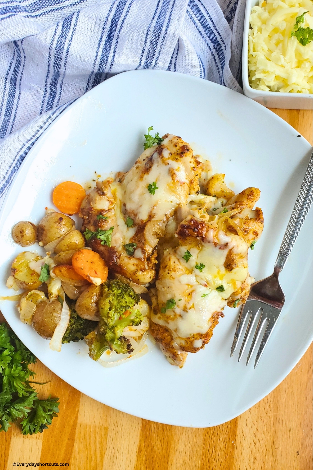 garlic parmesan chicken thighs on a plate with potatoes and veggies next to a fork, bowl of shredded parmesan cheese and blue and white stripped napkin