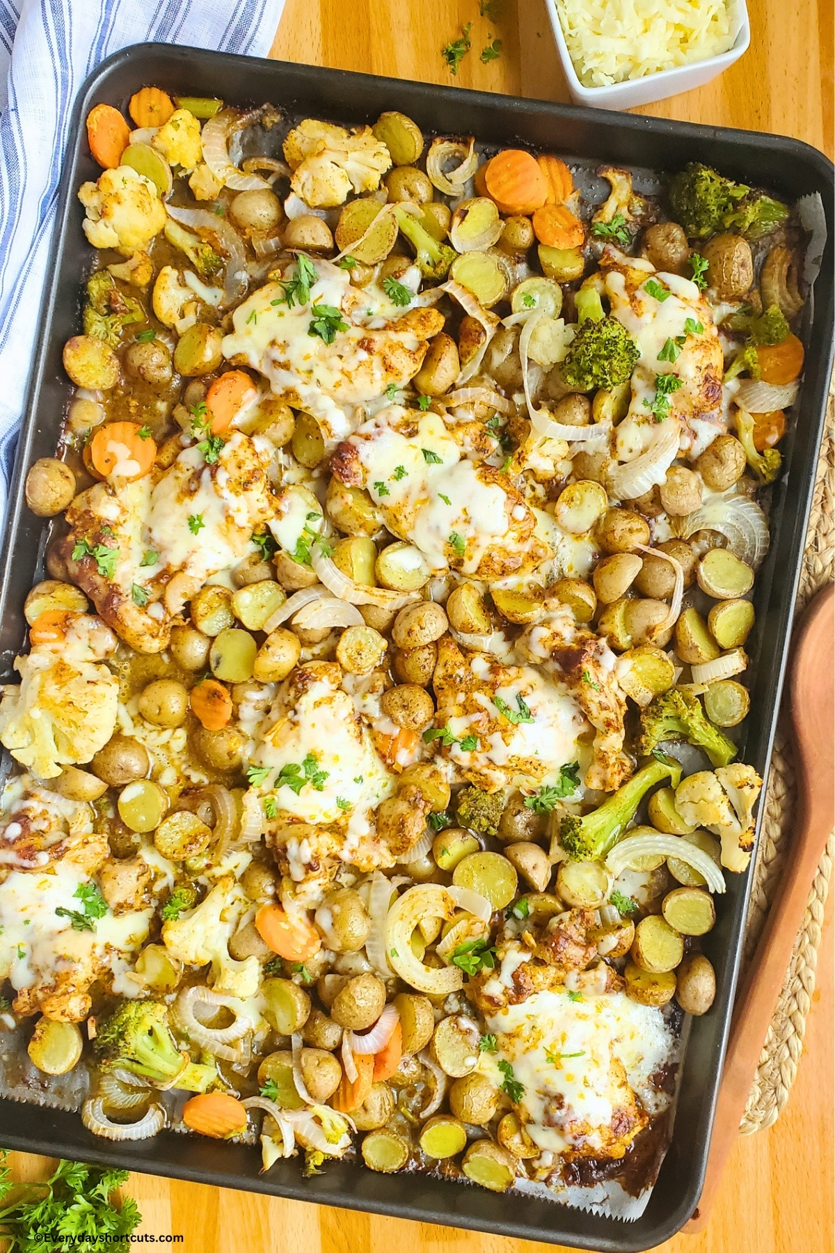 Garlic Parmesan Chicken Thighs with potatoes and veggies on a sheet pan next to a wooden spoon and blue and white striped napkin