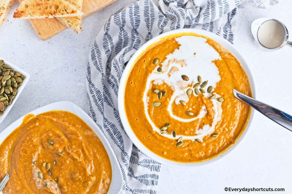 2 white bowls of creamy pumpkin soup next to a blue and white napkin, a bowl of cream and bowl of pumpkin seed