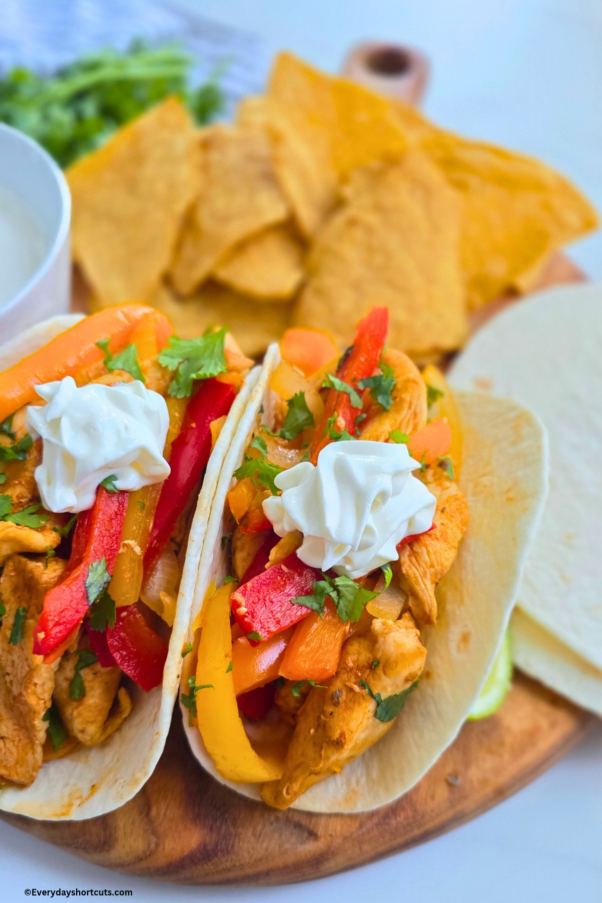 chicken fajitas with a dollop of sour cream and fresh cilantro on top on a wood board next to flour tortillas and tortilla chips