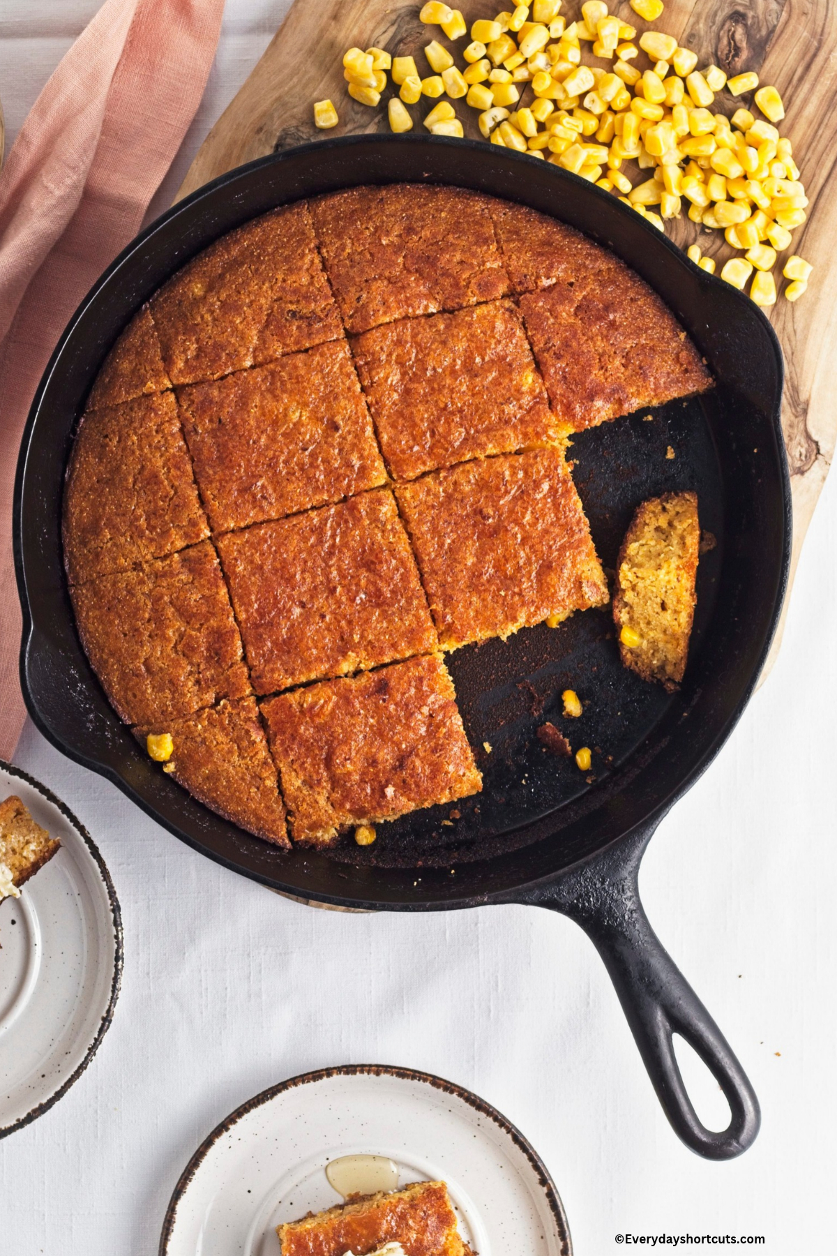 overhead shot of cornbread cut up in squares in a cast iron skillet next to corn kernels on a wood board